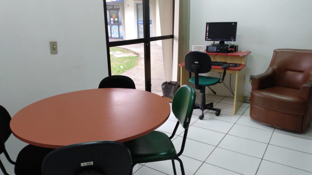 Sala onde aparecem mesa redonda com 4 cadeira. Mesa com computador e cadeira verde. Poltrona marrom de um lugar