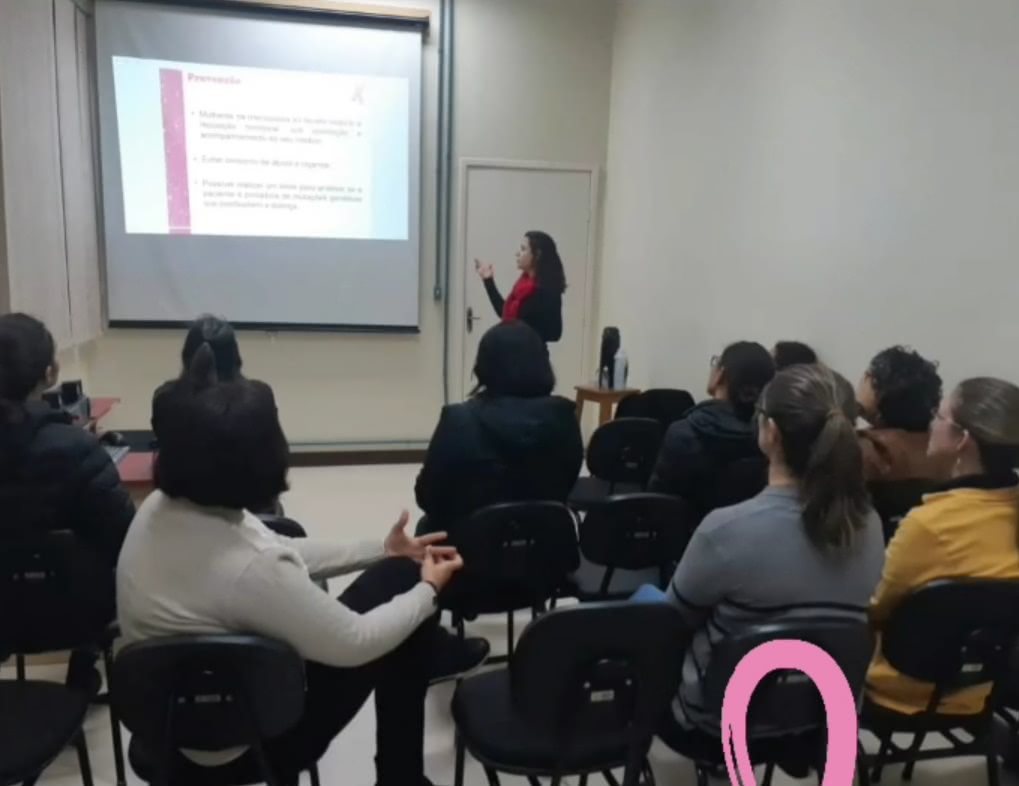 Uma imagem na sala de aula, aluna da enfermagem olhando para apresenação projetada na parede. Os ouvintes aparecem sentados nas cadeiras. todos estão de costas para camera e de frente para apresentação.