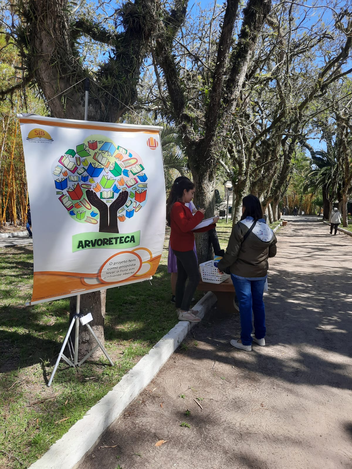 Fundo com árvores da praça Tamandaré, do lado esquerdo o banner do projeto arvoreteca e ao lado pessoas olhando alguns livros.