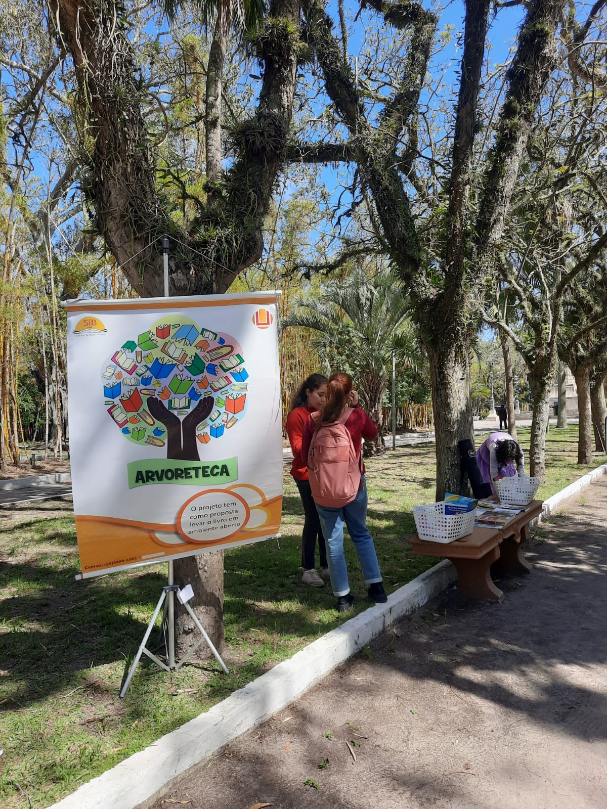 Fundo com árvores da praça Tamandaré, do lado esquerdo o banner do projeto arvoreteca e ao lado pessoas olhando alguns livros.