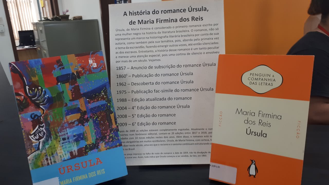 prateleira com dois livros posicionados em vertical, ao centro um informativo sobre a Históira do romance úrsula de Maria Firmina dos Reis. Livro da esquerda da foto capa colorida, abaixo barra azul clara escrito úrsula autora maria firmina reis. Livro da esquerda duas barras  de cor laranja superior e inferior ao centro barra branca escrito: maria firmina dos reis Úrsula. ao centro da barra superior laranja encontra-se um balão desenho branco com o nome da editora: Penguim e companhia das letras