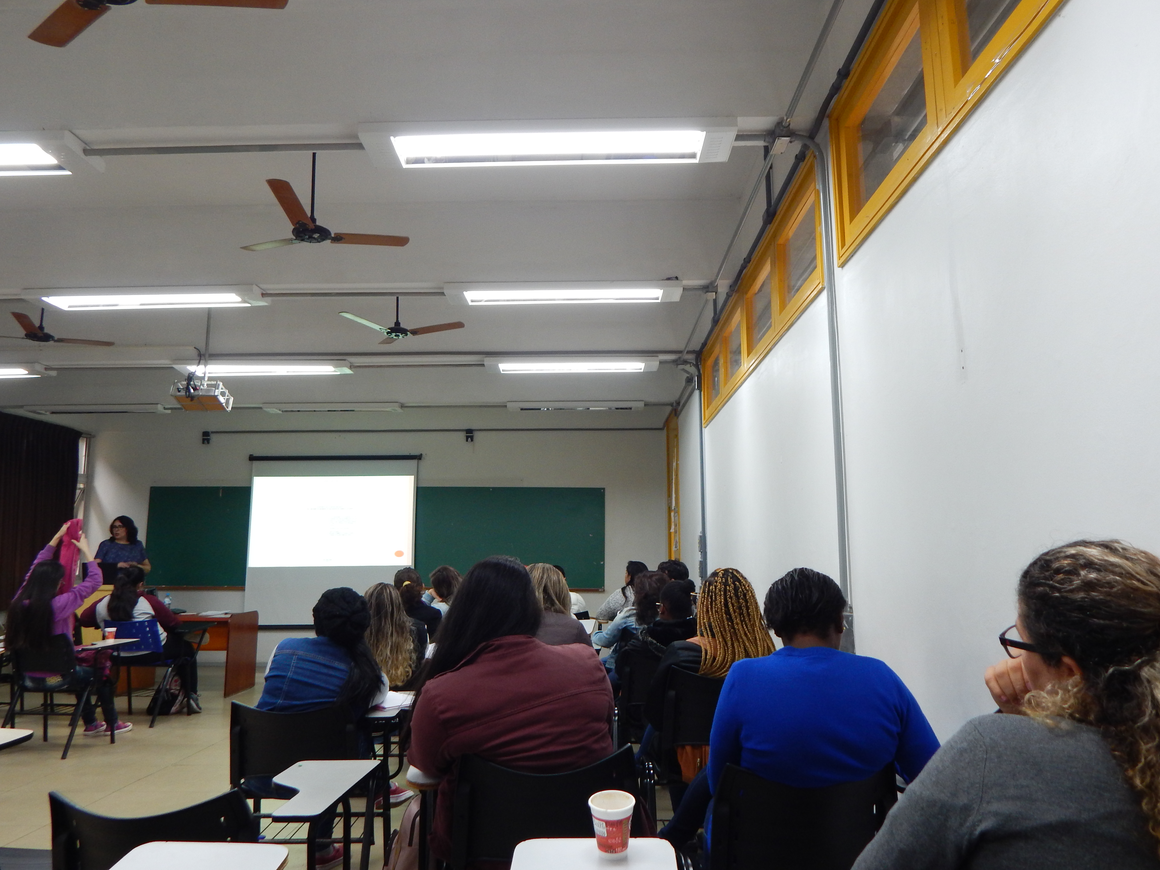 alunos sentados em sala de aula. Foram fotografados de costas olhando para a apresentaação em power point. A esquerda da foto posicionada em pé a Bibliotecária que está apresentando o treinamento