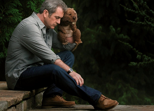Imagem com um fundo de árvores e a frente um homem de calça jeans e camisa sentado em um degrau segurando um urso de pelúcia na cor marrom.