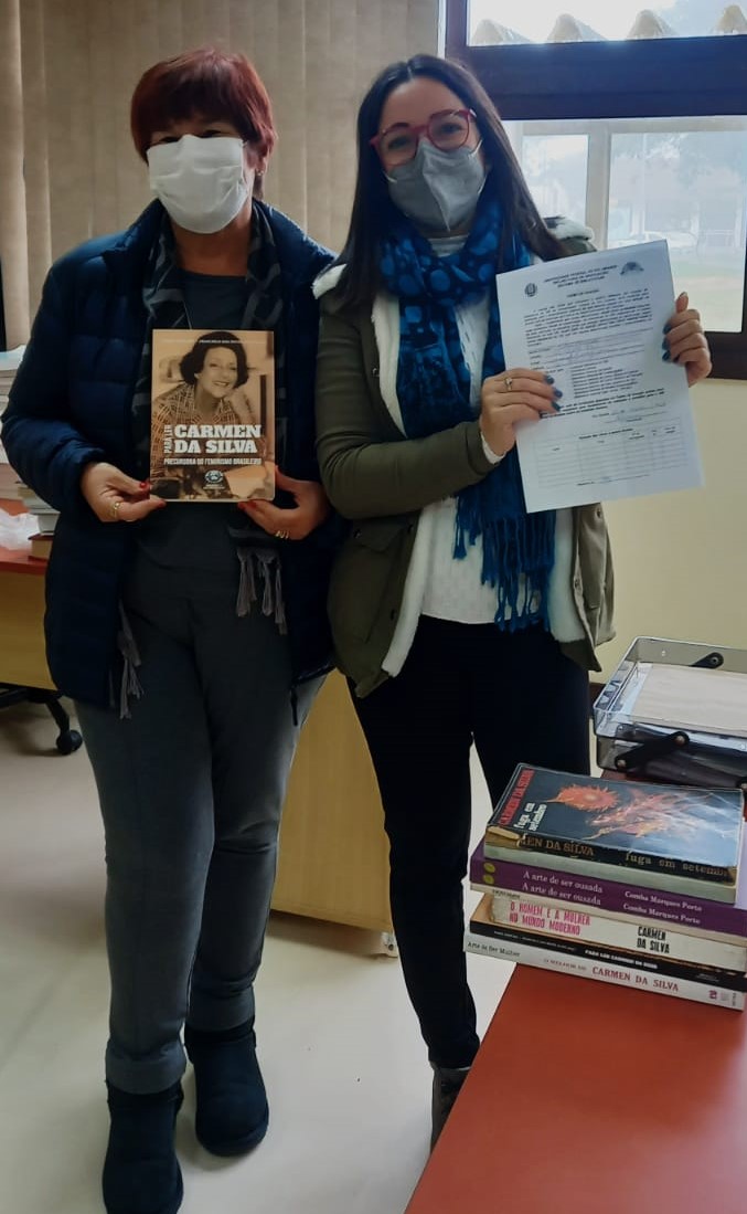 foto duas mulheres usando mascaras, a primeira segura o Livro da Carmen da Silva, a segunda segura o termo de doação