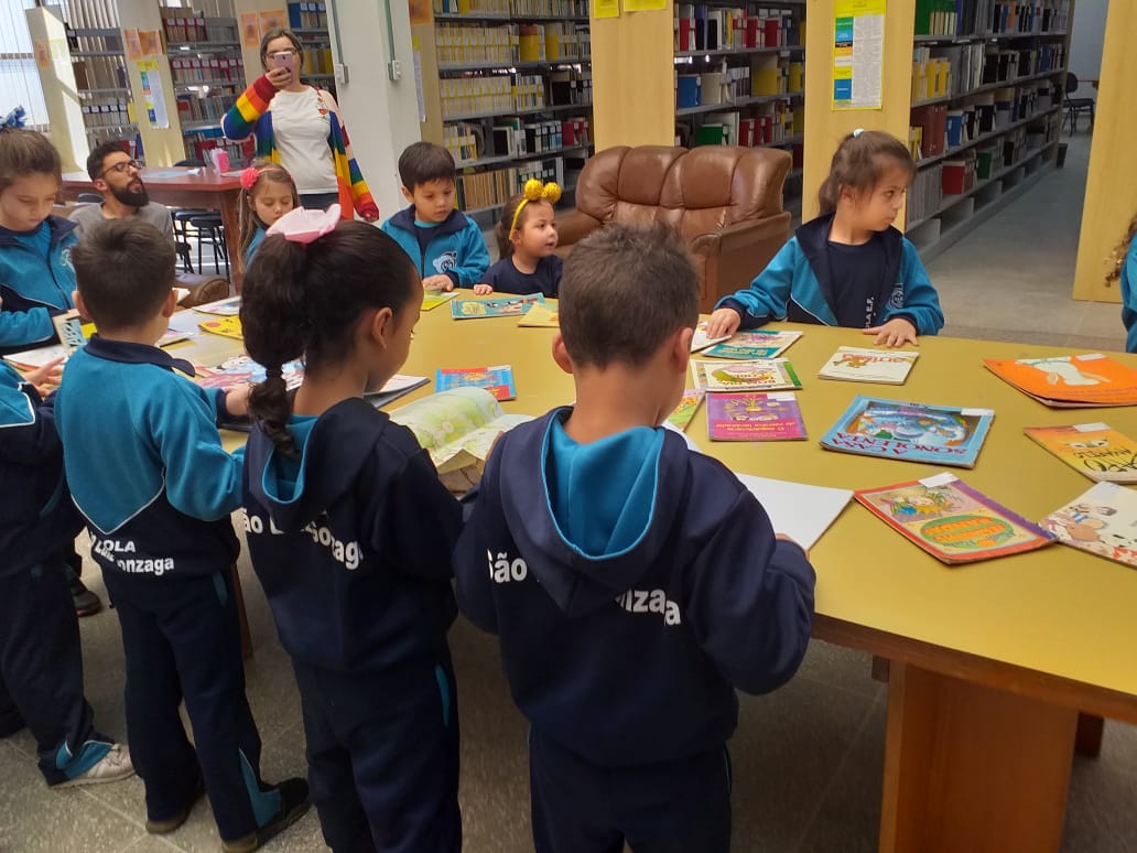 na imagem encontra-se uma mesa grande amarela no centro da biblioteca central  com alguns livros posicionados ao centro, as alunos da pré escola aparecem ao redor da mesa com seu uniformes em azul claro e escuro