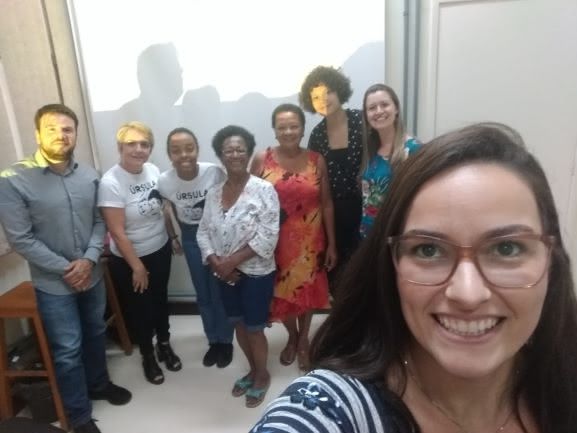 Bibliotecária faz selfie: no canto inferior direito o perfil da biblitecária, ela usa óculos, expressa sorriso e tem cabelos castanhos longos. Ao fundo da direita para esquerda: bibliotecário usa camisa cinza com botões calça jeans, tem barba, esta com as mãos cruzadas para frente. Logo a direita duas palestrantes utilizam camisetas brancas escrito Ursula, ambas expessam sorrisos. A seguilr uma senhora usando óculos camisa branca, berbuda jeans, está com as mãos cruzadas a frente, ao lado dela uma mulher com vestido estampado longo. a Seguir uma mulher jovem cabelos curtos crespos, está vestindo blusa azul com bolinhas brancas, e por último mulher cabelos longos loiros. Todos expressam sorrisos