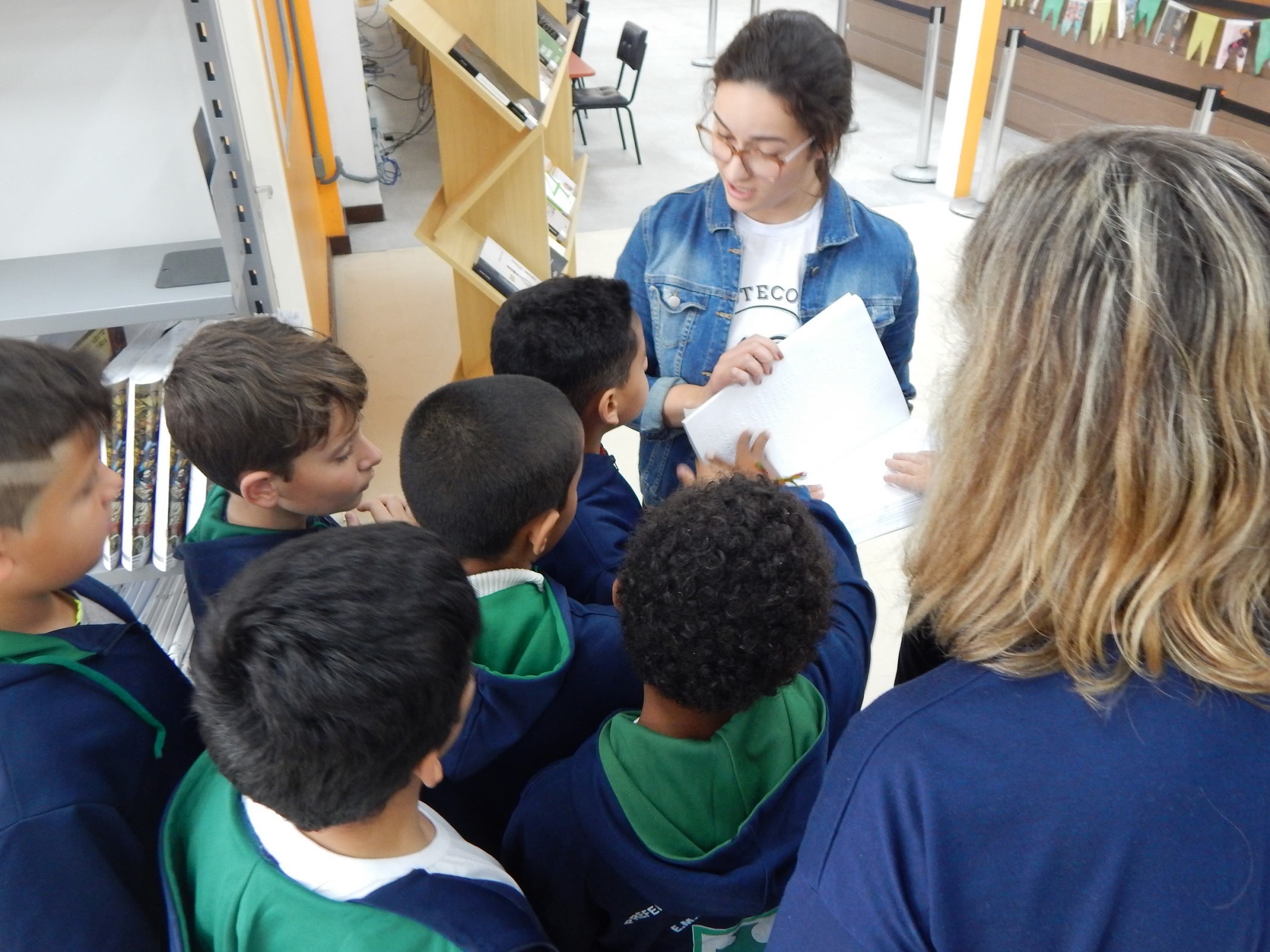 alunos aparecem de costas, usando uniforme azul e verde. A bibliotecária segura em suas mãos um livro em braille, os alunos escutam as explicações atentos