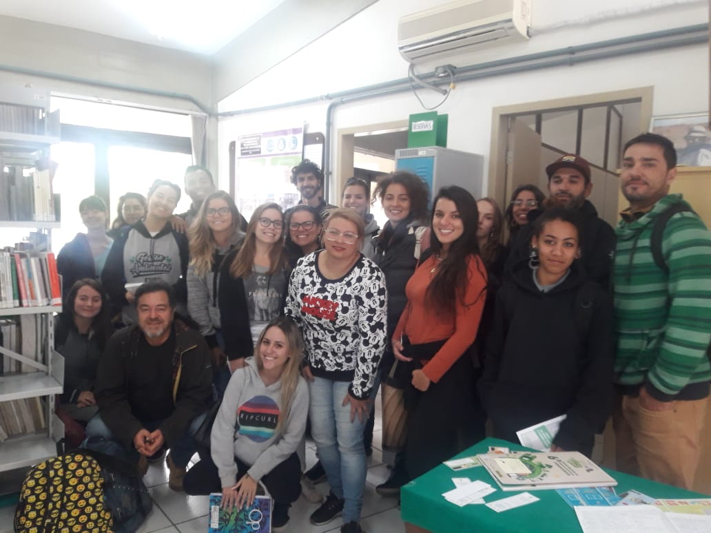 foto no interior da sala verde, onde os alunos aparecem agrupados em pé , e apenas 3 deles estão abaixados, a esquerda uma estante de livros, a frente uma mesa