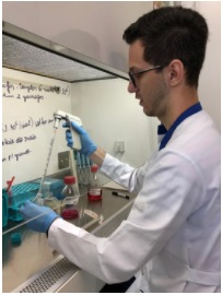pesquisador no laboratório, manipulando tubos de ensaio, usa luva azul, jaleco branco, óculos armação preta, foto de perfil