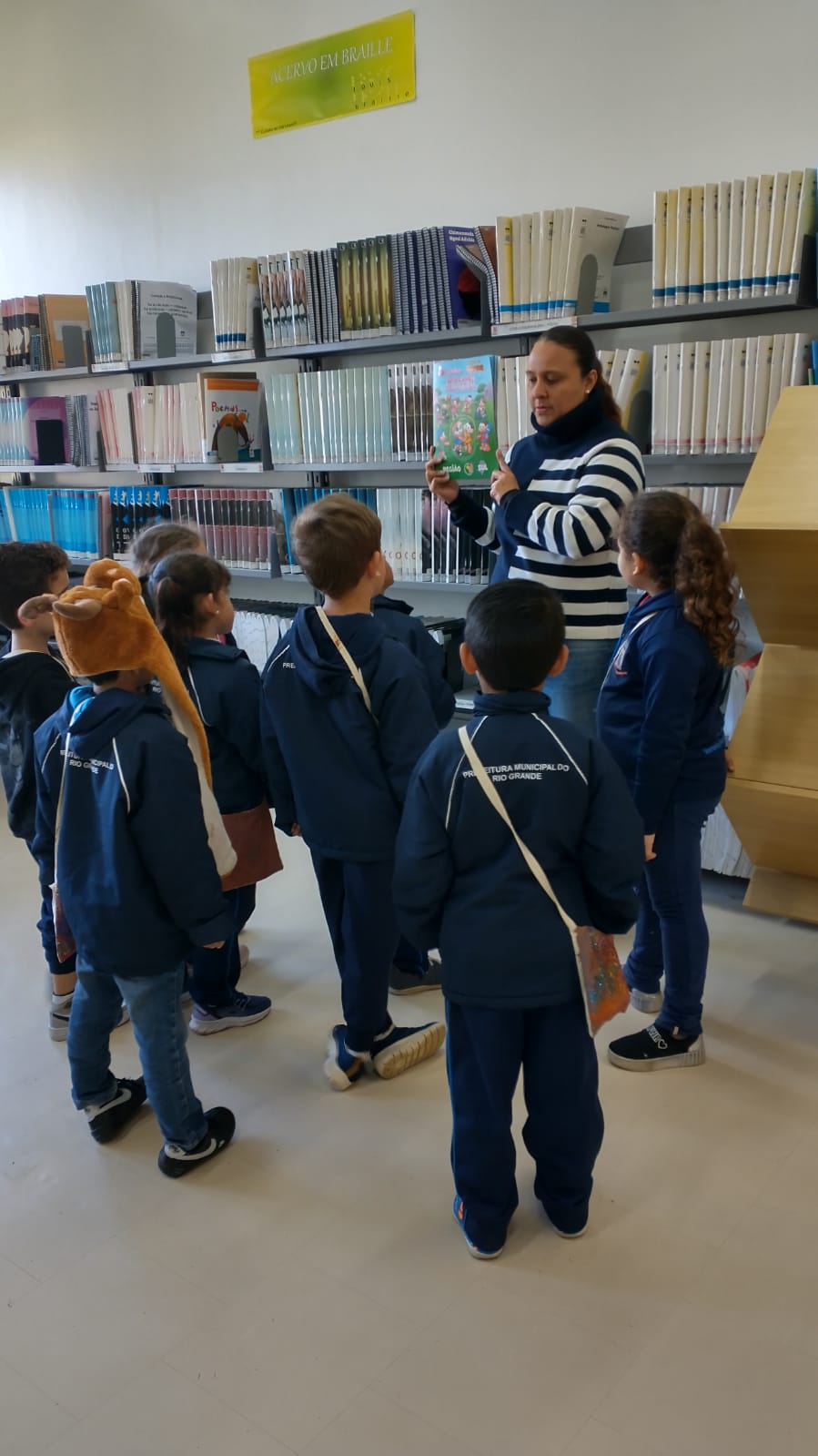 grupo de crianças   em volta de uma bibliotecaria que segura um livro nas mãos, dentro de uma biblioteca.